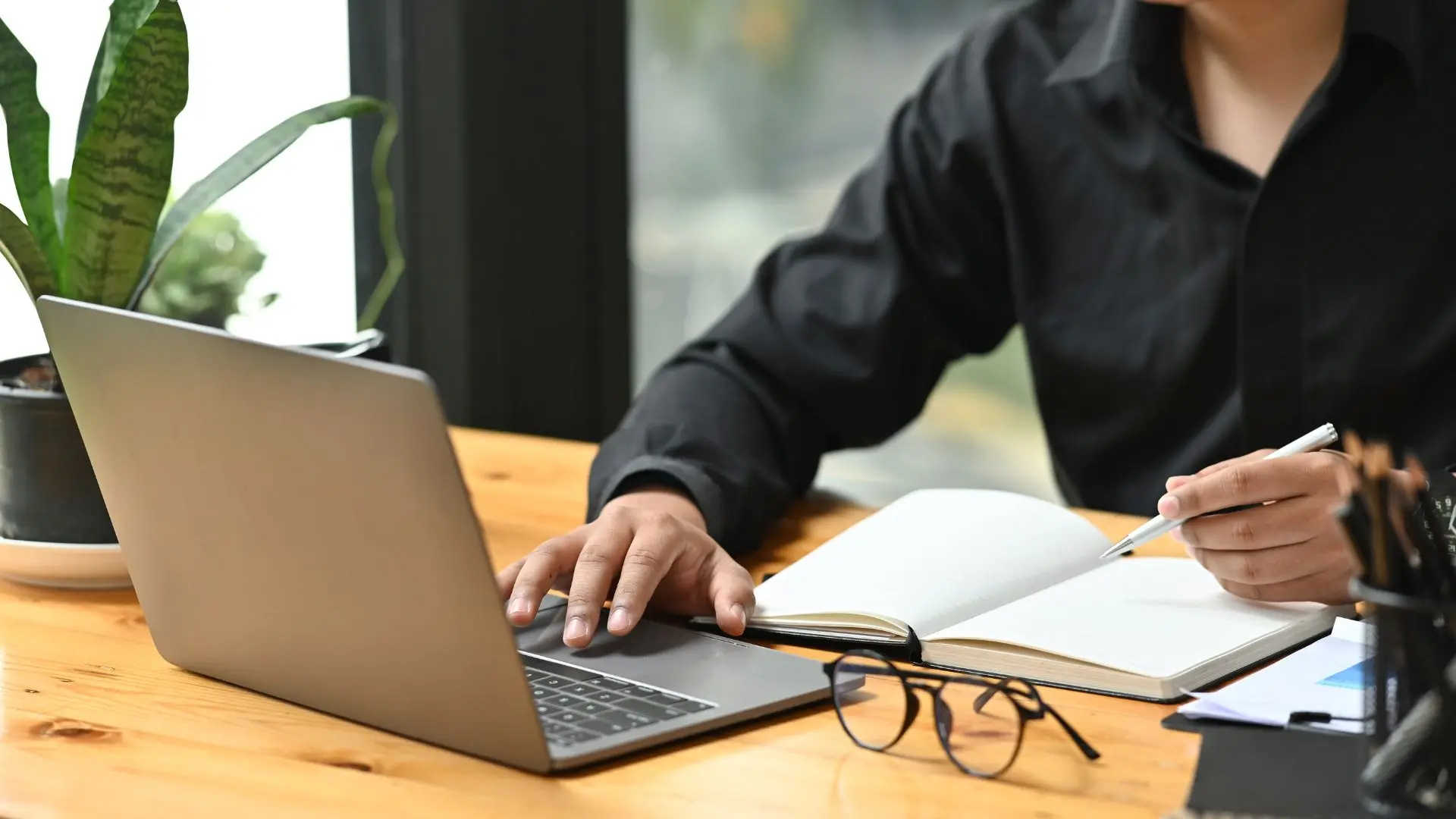 Hombre con camisa sentado en escritorio frente a un portátil y escribiendo en un cuaderno sobre servicios de copywriting