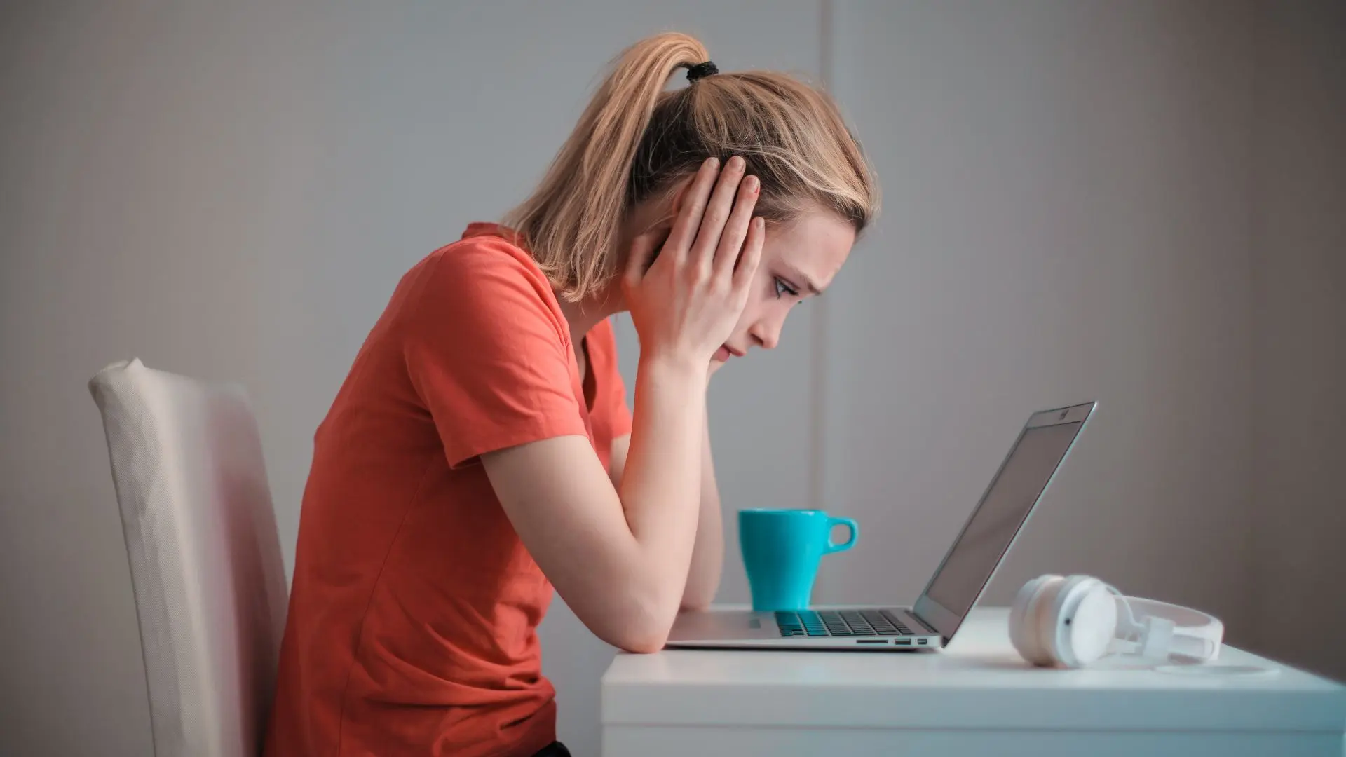 Mujer preocupada sobre errores de traducción sentada frente a una mesa en la que hay una taza y unos auriculares, mirando la pantalla de su portátil