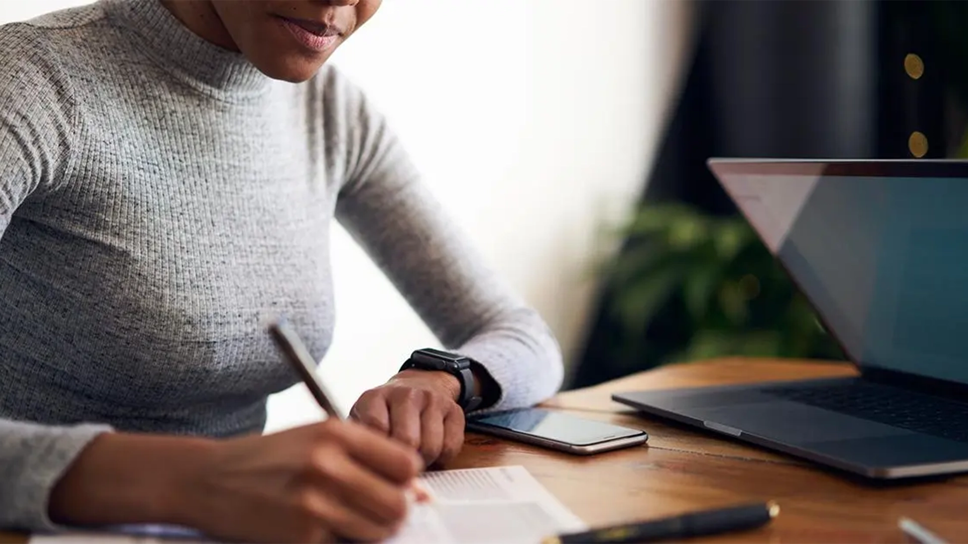 Mujer sentada escribiendo en una hoja sobre las principales dificultades de la traducción de publicidad con un portátil y un teléfono móvil sobre la mesa