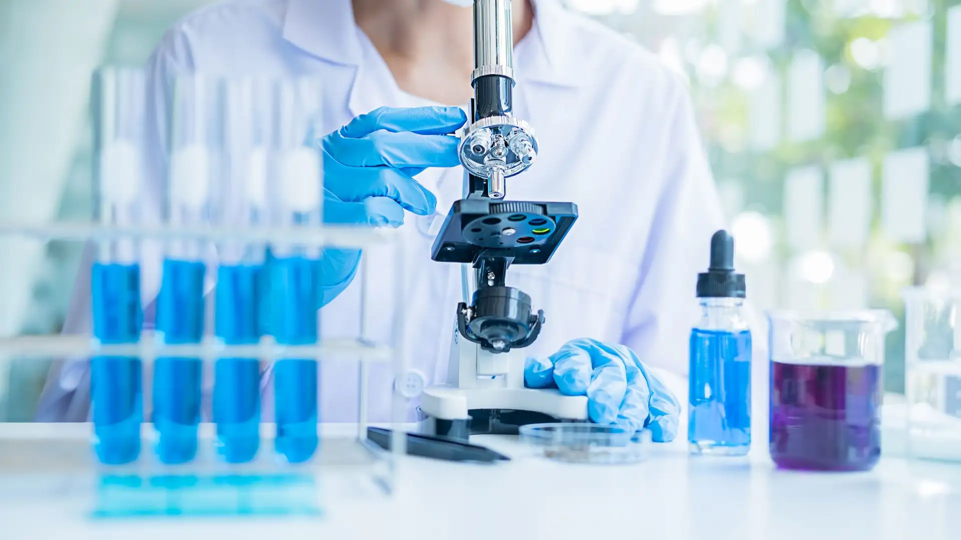 Mujer con bata y guantes utilizando un microscopio en una mesa sobre la que hay probetas y botes sobre traducción farmacéutica
