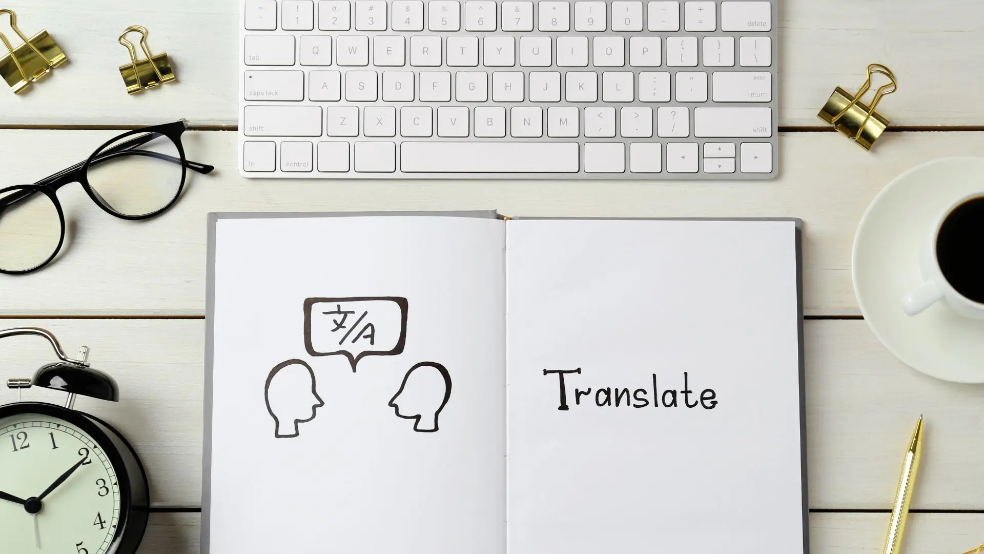 En un escritorio hay un teclado de ordenador, unas gafas, un reloj, una taza de café y una libreta con símbolos de traducción de contenidos
